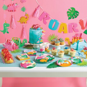 Puede incluir: Una mesa de fiesta con tem&aacute;tica de dinosaurios con platos, servilletas y vasos de papel azul y verde. Hay un pastel azul con chispas rosas y verdes, cupcakes con chispas de colores y una caja de pastel con forma de dinosaurio. La mesa est&aacute; decorada con una pancarta de dinosaurio rosa y verde que dice "ROAR!"