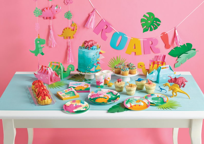 Puede incluir: Una mesa de fiesta con tem&aacute;tica de dinosaurios con platos, servilletas y vasos de papel azul y verde. Hay un pastel azul con chispas rosas y verdes, cupcakes con chispas de colores y una caja de pastel con forma de dinosaurio. La mesa est&aacute; decorada con una pancarta de dinosaurio rosa y verde que dice "ROAR!"