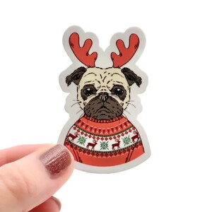 May include: A beige pug wearing a red and green Christmas sweater with reindeer and snowflakes. The pug has antlers on its head.