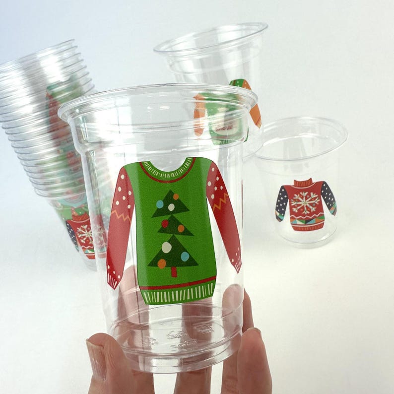 May include: Clear plastic cups with a festive Christmas sweater design. The sweaters are green with a Christmas tree, red sleeves, and white polka dots. Other cups have a red sweater design with a snowflake.