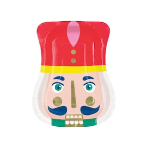 Puede incluir: Un plato de papel blanco con un diseño de cascanueces rojo, amarillo y azul. El cascanueces tiene un sombrero rosa, botones dorados y una barba verde.