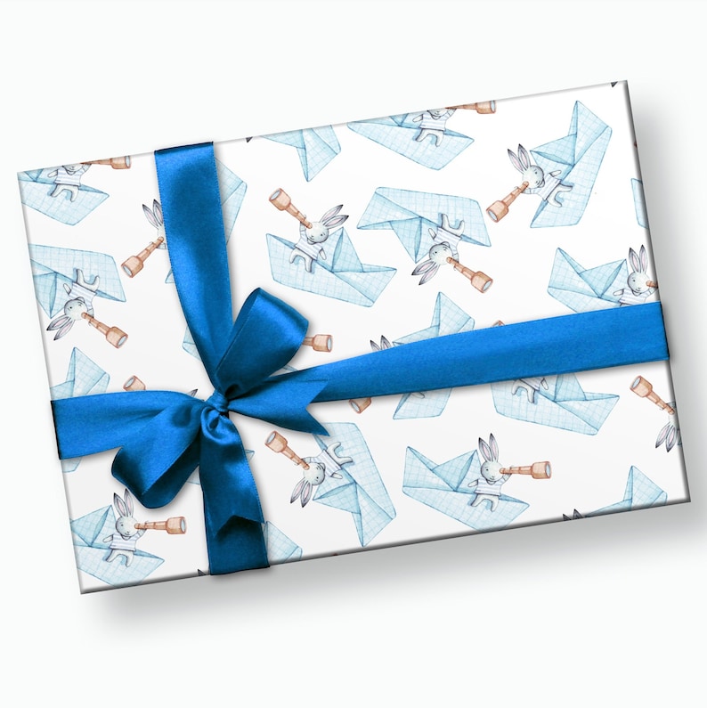 Puede incluir: Una caja de regalo envuelta en papel con un patr&oacute;n de barcos de papel azules y conejos con telescopios sobre un fondo blanco. La caja est&aacute; atada con una cinta y un lazo de sat&eacute;n azul brillante, adecuada para cualquier celebraci&oacute;n.