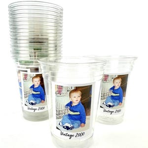 Puede incluir: Vasos de pl&aacute;stico transparente con una foto de beb&eacute; y el texto "Vintage 2000". Los vasos est&aacute;n apilados y dispuestos sobre una superficie blanca. El beb&eacute; lleva una camisa azul y vaqueros.