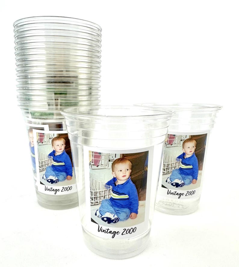 Puede incluir: Vasos de pl&aacute;stico transparente con una foto de beb&eacute; y el texto "Vintage 2000". Los vasos est&aacute;n apilados y dispuestos sobre una superficie blanca. El beb&eacute; lleva una camisa azul y vaqueros.
