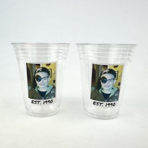 May include: Two clear plastic cups each feature a photo of a person wearing a pirate hat and eye patch. The photo is printed on a sticker with the text "EST. 1990" below the image. The cups are on a white surface.