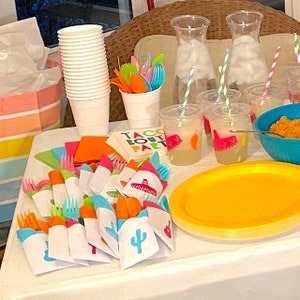 May include: A table setting for a taco party with colorful napkins, cutlery, and cups. The table is decorated with a yellow and orange tablecloth and a sign that says "Taco Bout a Party".