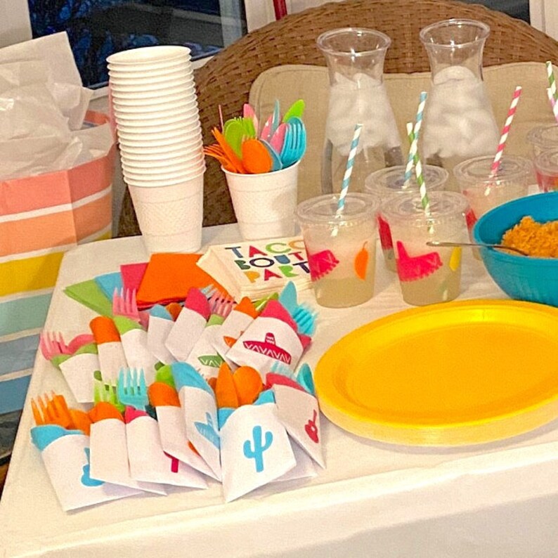 May include: A table setting for a taco party with colorful napkins, cutlery, and cups. The table is decorated with a yellow and orange tablecloth and a sign that says "Taco Bout a Party".