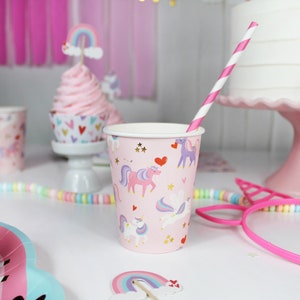 May include: Pink and white paper cups with a unicorn and heart design. The cups are decorated with gold stars and glitter. A pink and white striped straw is in one of the cups.