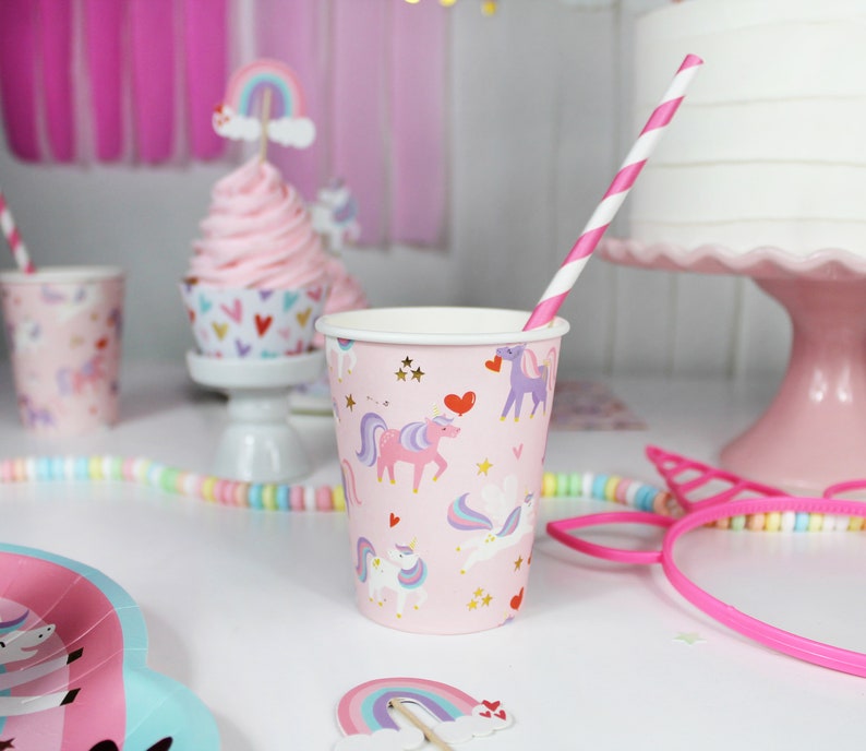 May include: Pink and white paper cups with a unicorn and heart design. The cups are decorated with gold stars and glitter. A pink and white striped straw is in one of the cups.