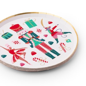 May include: White paper plates with a gold rim and a festive Nutcracker design. The plates feature illustrations of a Nutcracker soldier, a ballerina, and other holiday-themed graphics in red, green, and pink.