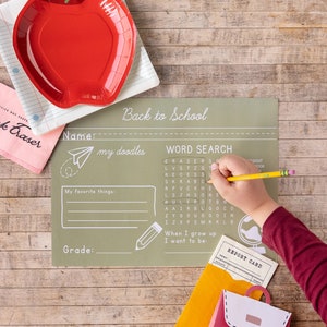 May include: A red apple-shaped plate sits on a notebook paper plate. A child's hand is writing on a green Back to School placemat with a word search. A pink eraser and a backpack-shaped card are also visible.