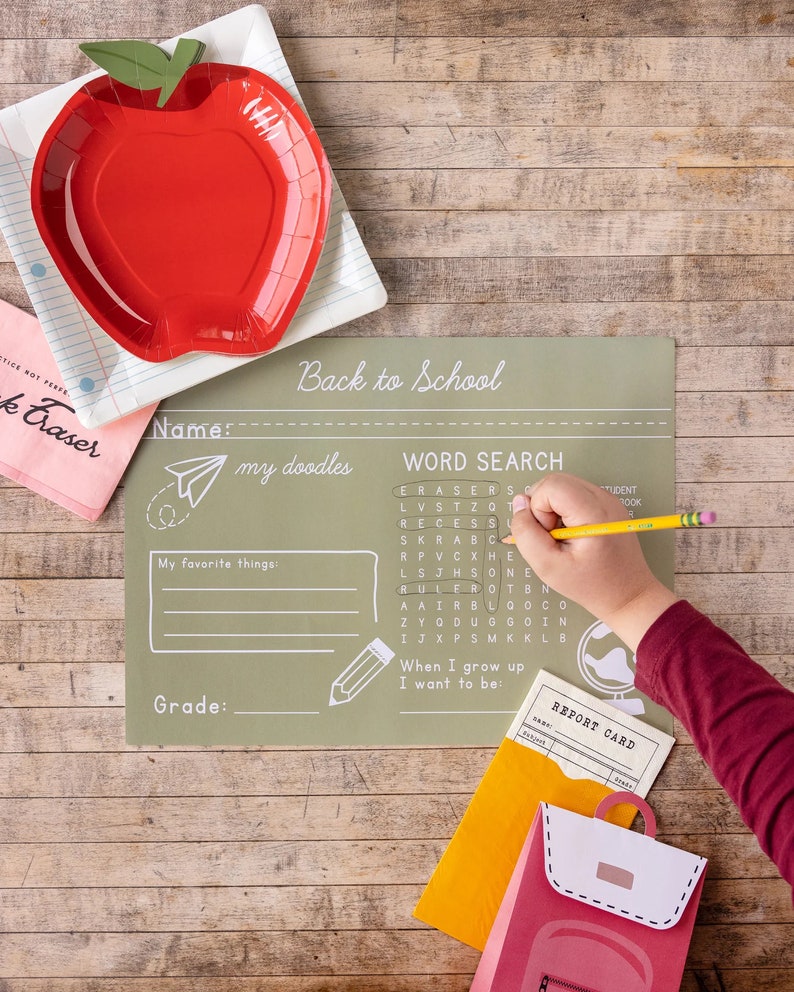 May include: A red apple-shaped plate sits on a notebook paper plate. A child's hand is writing on a green Back to School placemat with a word search. A pink eraser and a backpack-shaped card are also visible.