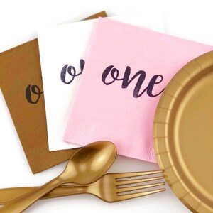 May include: A set of party supplies for a first birthday party, including a gold paper plate, gold plastic fork and spoon, and three napkins with the word "one" written on them in black ink. The napkins are brown, white, and pink.