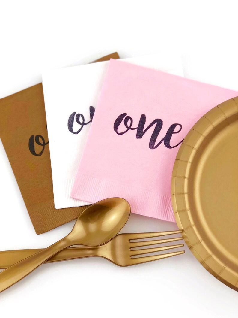May include: A set of party supplies for a first birthday party, including a gold paper plate, gold plastic fork and spoon, and three napkins with the word "one" written on them in black ink. The napkins are brown, white, and pink.