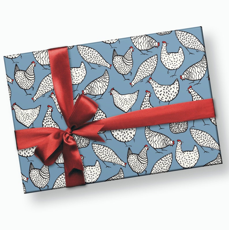 K&ouml;nnte beinhalten: Blaues Geschenkpapier mit einem sich wiederholenden Muster aus wei&szlig;en H&uuml;hnern mit schwarzen Markierungen. Die H&uuml;hner haben rote K&auml;mme und Kehllappen. Ein rotes Band ist mit einer Schleife um die Schachtel gebunden.