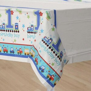 May include: White tablecloth with a blue border and a repeating pattern of trains, cars, and the number 1. The words "birthday boy" are printed on the tablecloth. The design includes stars and a helicopter.