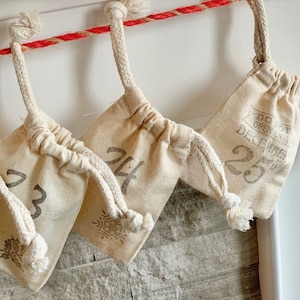 Puede incluir: Calendario de Adviento con peque&ntilde;as bolsas de tela color crema colgadas de una cuerda roja y blanca. Cada bolsa est&aacute; numerada con una fuente gris, algunas con dise&ntilde;os de copos de nieve o hojas. Una bolsa dice "DO NOT OPEN UNTIL DECEMBER 25."