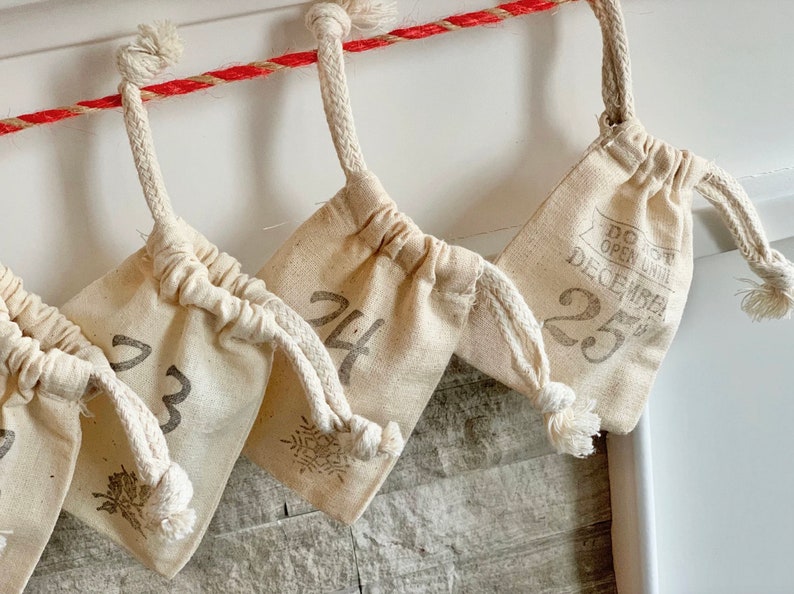 Puede incluir: Calendario de Adviento con peque&ntilde;as bolsas de tela color crema colgadas de una cuerda roja y blanca. Cada bolsa est&aacute; numerada con una fuente gris, algunas con dise&ntilde;os de copos de nieve o hojas. Una bolsa dice "DO NOT OPEN UNTIL DECEMBER 25."