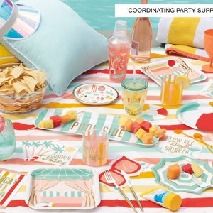 May include: A colorful summer party table setting with a striped tablecloth, paper plates, napkins, and cups with a tropical theme. The table is decorated with a pineapple, sunglasses, and a beach towel. The text on the napkins reads "Low Key Day Drinker", "No Shade", "Vacay All Day", and "Pool Side".