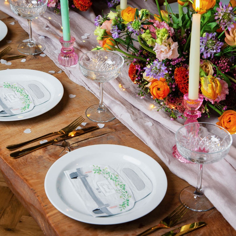 May include: A wedding table setting with white plates, gold silverware, and clear glasses. The centerpiece is a bouquet of colorful flowers with pink candlesticks and white napkins with a car design and the text "Just Married".