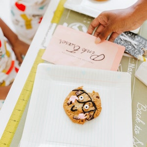 May include: A decorated chocolate chip cookie with a face design on a white square plate. The cookie has a bite taken out of it and is decorated with black icing and pink frosting. A pink card with the text "Pink Fraser" is in the background.