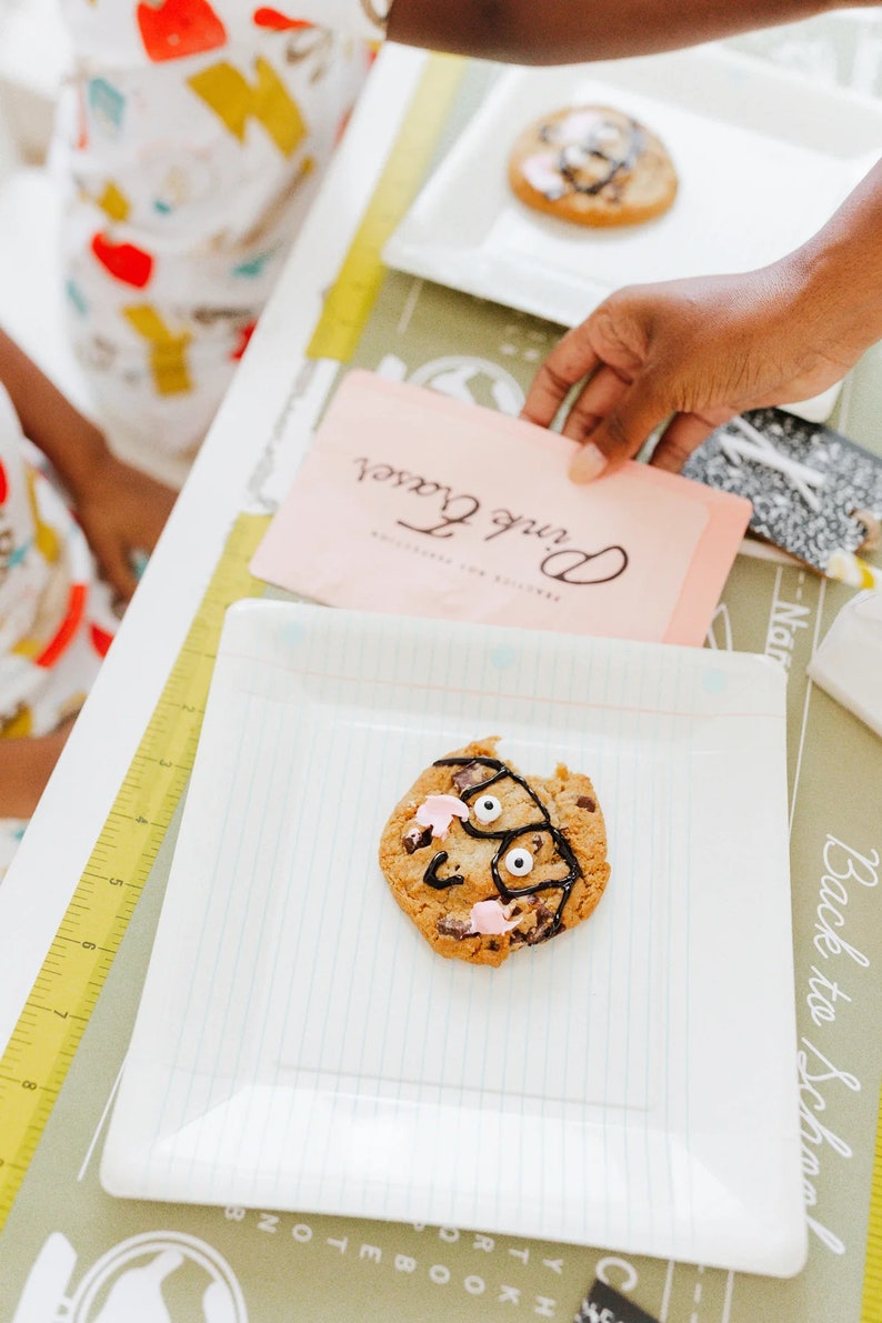 May include: A decorated chocolate chip cookie with a face design on a white square plate. The cookie has a bite taken out of it and is decorated with black icing and pink frosting. A pink card with the text "Pink Fraser" is in the background.