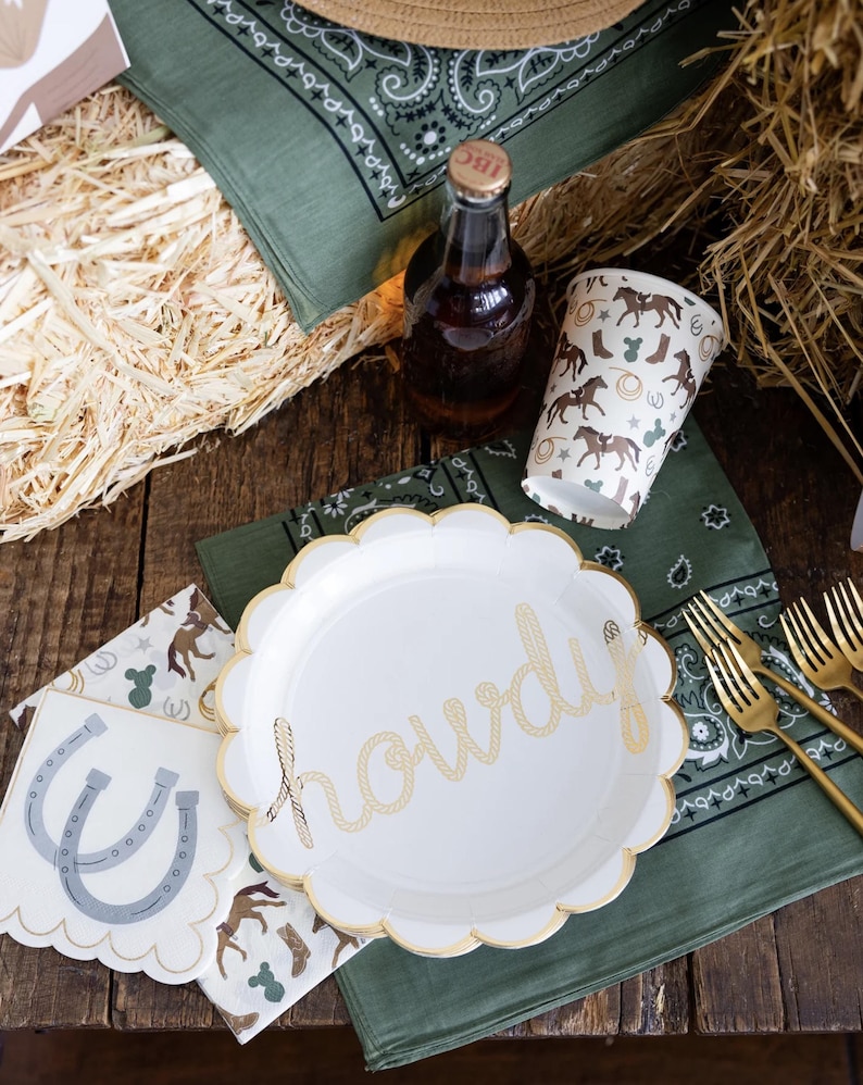 May include: A rustic party setting with a white plate featuring the word "howdy" in gold rope lettering. The scene includes a bottle, paper cups, napkins, and gold forks, all with a cowboy theme. Green bandanas and straw complete the western aesthetic.
