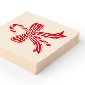 Puede incluir: Una pila de servilletas de papel blanquecino. Cada servilleta presenta un diseño de bastón de caramelo y lazo rojo y blanco. El lazo tiene rayas rojas y está atado en el centro. Las servilletas están apiladas ordenadamente.