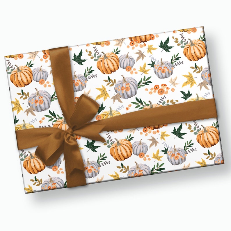 May include: A white gift box with a brown ribbon bow. The box is wrapped in paper with a pattern of orange and gray pumpkins, green leaves, and yellow flowers.