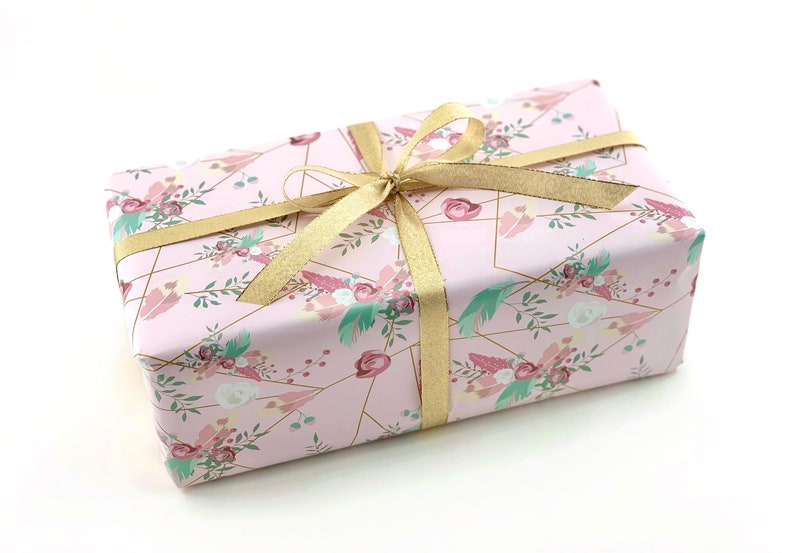 K&ouml;nnte beinhalten: Eine rechteckige Geschenkbox, verpackt in rosa floral gemustertem Papier mit goldenen geometrischen Linien. Die Box ist mit einem goldenen Band zu einer Schleife gebunden. Das Blumenmuster umfasst rosa, wei&szlig;e und t&uuml;rkisfarbene Blumen und Federn.