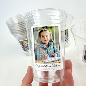 May include: Clear plastic cups with a photo of a young girl and the text "Congratulations Madison CLASS OF 2025". The cups are stacked and held by a hand.