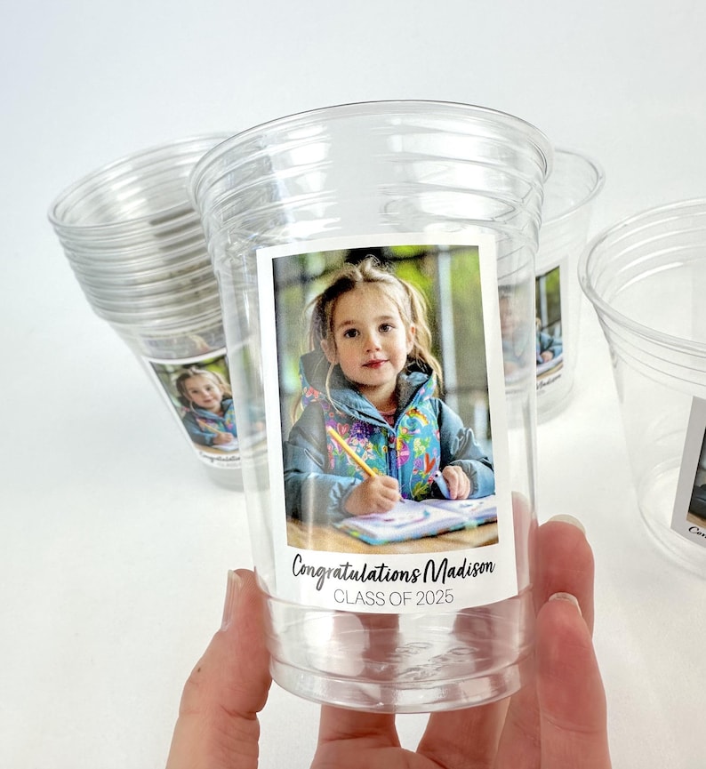 May include: Clear plastic cups with a photo of a young girl and the text "Congratulations Madison CLASS OF 2025". The cups are stacked and held by a hand.