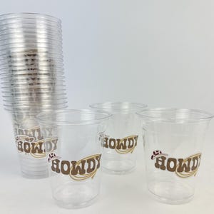 Puede incluir: Vasos de pl&aacute;stico transparente con un dise&ntilde;o "HOWDY" marr&oacute;n y dorado y un sombrero de vaquero. Una pila de vasos a la izquierda, con tres vasos individuales delante. Adecuados para fiestas.