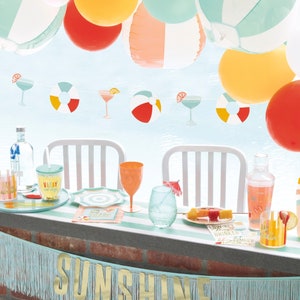 May include: A turquoise and gold banner with the word "Sunshine" on it hangs above a table with a variety of summer-themed party supplies, including colorful balloons, cocktail glasses, and a cocktail shaker.