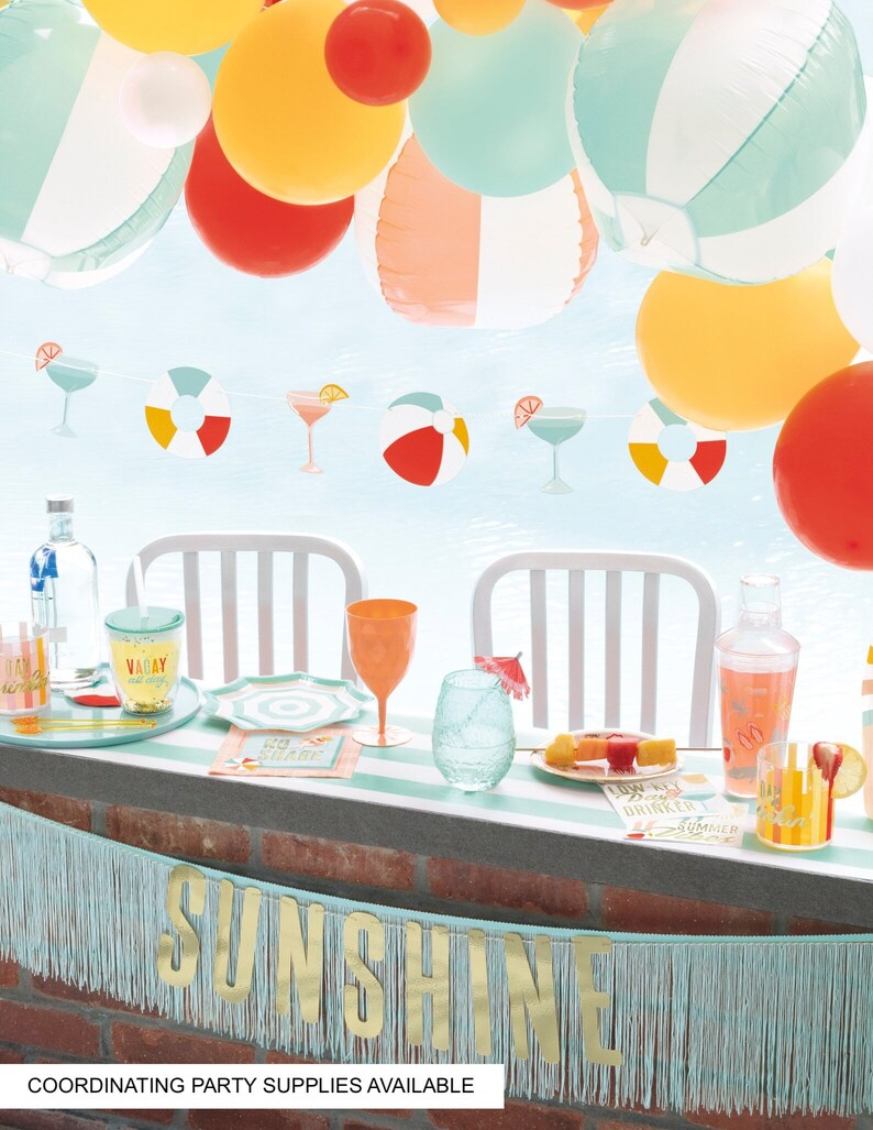 May include: A turquoise and gold banner with the word "Sunshine" on it hangs above a table with a variety of summer-themed party supplies, including colorful balloons, cocktail glasses, and a cocktail shaker.