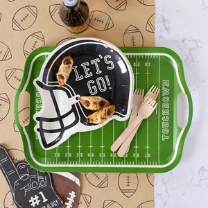 May include: A football-themed party setup featuring a green tray designed like a football field, a black helmet-shaped plate with "LET'S GO!" text, and wooden forks. Includes a foam finger, football-shaped decor, and a brown paper with football illustrations.