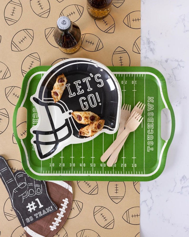 May include: A football-themed party setup featuring a green tray designed like a football field, a black helmet-shaped plate with "LET'S GO!" text, and wooden forks. Includes a foam finger, football-shaped decor, and a brown paper with football illustrations.