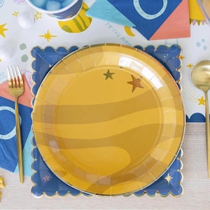 Peut inclure: Une assiette en papier jaune avec un motif de planète et des étoiles dorées. L'assiette est placée sur une assiette en papier bleue avec un bord festonné.