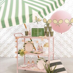 Puede incluir: Un carrito rosa exhibe decoraciones de fiesta con tem&aacute;tica de tenis. El carrito contiene platos, vasos y botellas de l&iacute;quido rosa. Un gran globo rosa con recortes de raquetas de tenis flota cerca. Una sombrilla a rayas verdes y blancas proporciona sombra.
