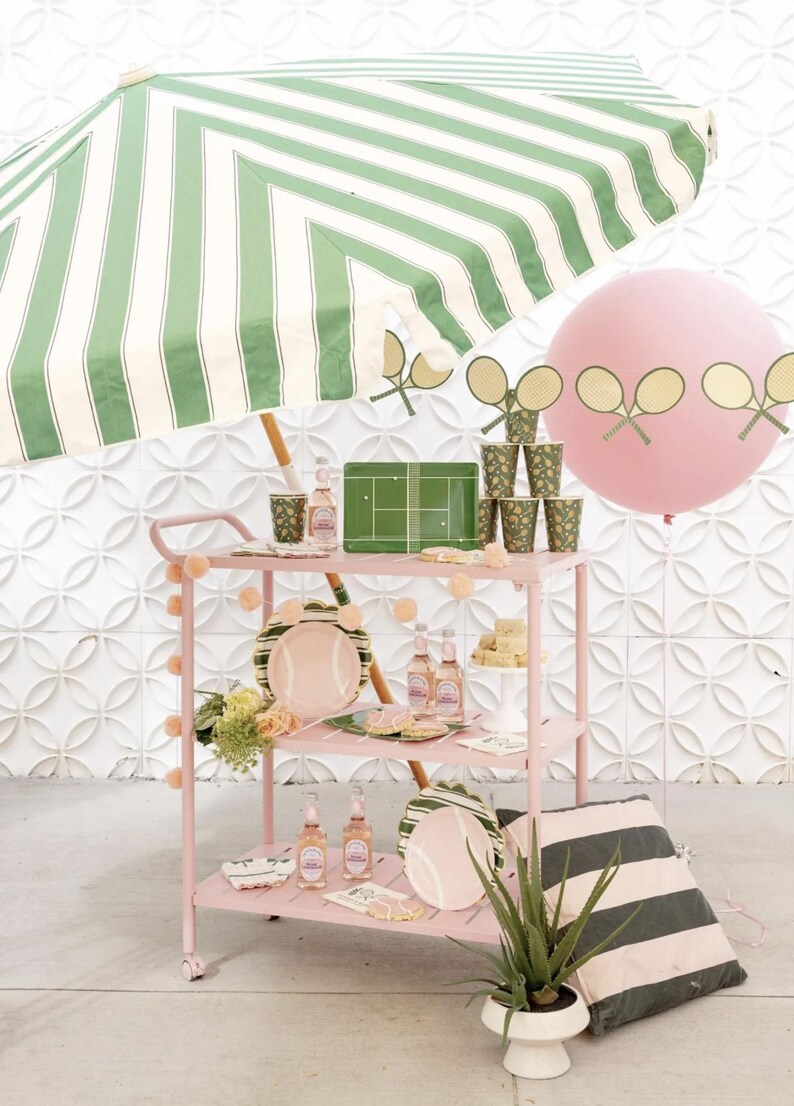 Puede incluir: Un carrito rosa exhibe decoraciones de fiesta con tem&aacute;tica de tenis. El carrito contiene platos, vasos y botellas de l&iacute;quido rosa. Un gran globo rosa con recortes de raquetas de tenis flota cerca. Una sombrilla a rayas verdes y blancas proporciona sombra.