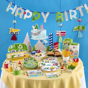 May include: A birthday party table setting with a blue and green "Happy Birthday" banner, a blue and white cake with a number 6 on top, and a variety of party supplies with a transportation theme. The table is covered in a yellow tablecloth and features plates, napkins, cups, and other decorations with cars, trucks, and airplanes.