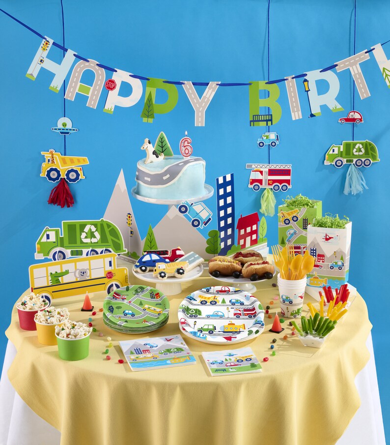 May include: A birthday party table setting with a blue and green "Happy Birthday" banner, a blue and white cake with a number 6 on top, and a variety of party supplies with a transportation theme. The table is covered in a yellow tablecloth and features plates, napkins, cups, and other decorations with cars, trucks, and airplanes.