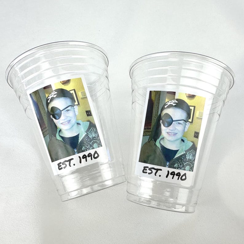 May include: Two clear plastic cups with a photo of a person wearing a pirate hat and eye patch. The photo has the text "EST. 1990" at the bottom. The cups are on a white surface.