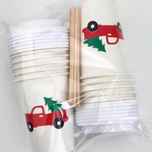 May include: A set of disposable paper cups with lids and wooden stirrers, packaged in a clear plastic bag. The cups are cream-colored and feature a red truck graphic carrying a green Christmas tree. The lids are white.