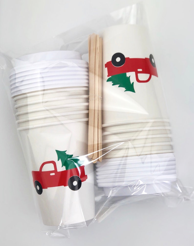 May include: A set of disposable paper cups with lids and wooden stirrers, packaged in a clear plastic bag. The cups are cream-colored and feature a red truck graphic carrying a green Christmas tree. The lids are white.
