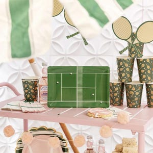 Puede incluir: Un carrito rosa exhibe art&iacute;culos para fiestas con tem&aacute;tica de tenis. Incluye una bandeja rectangular verde con dise&ntilde;o de cancha de tenis, vasos de papel con patrones de raquetas y platos rosas. Elementos decorativos: recortes de raquetas y pompones.