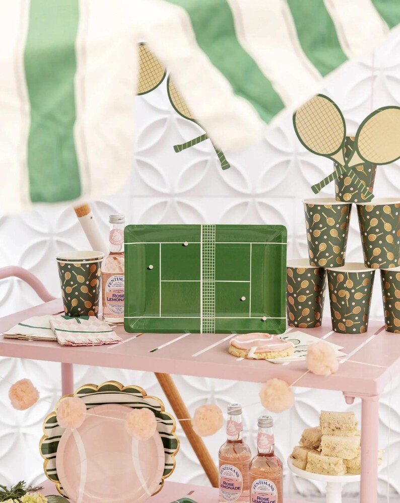 Puede incluir: Un carrito rosa exhibe art&iacute;culos para fiestas con tem&aacute;tica de tenis. Incluye una bandeja rectangular verde con dise&ntilde;o de cancha de tenis, vasos de papel con patrones de raquetas y platos rosas. Elementos decorativos: recortes de raquetas y pompones.