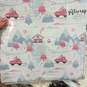 May include: A pack of three sheets of gift wrap featuring a winter wonderland scene with pink and blue houses, trees, and mountains. The wrapping paper is decorated with cars with dogs in them and the text "giftwrap boutique wrapping paper 3 sheets - 30" x 20".