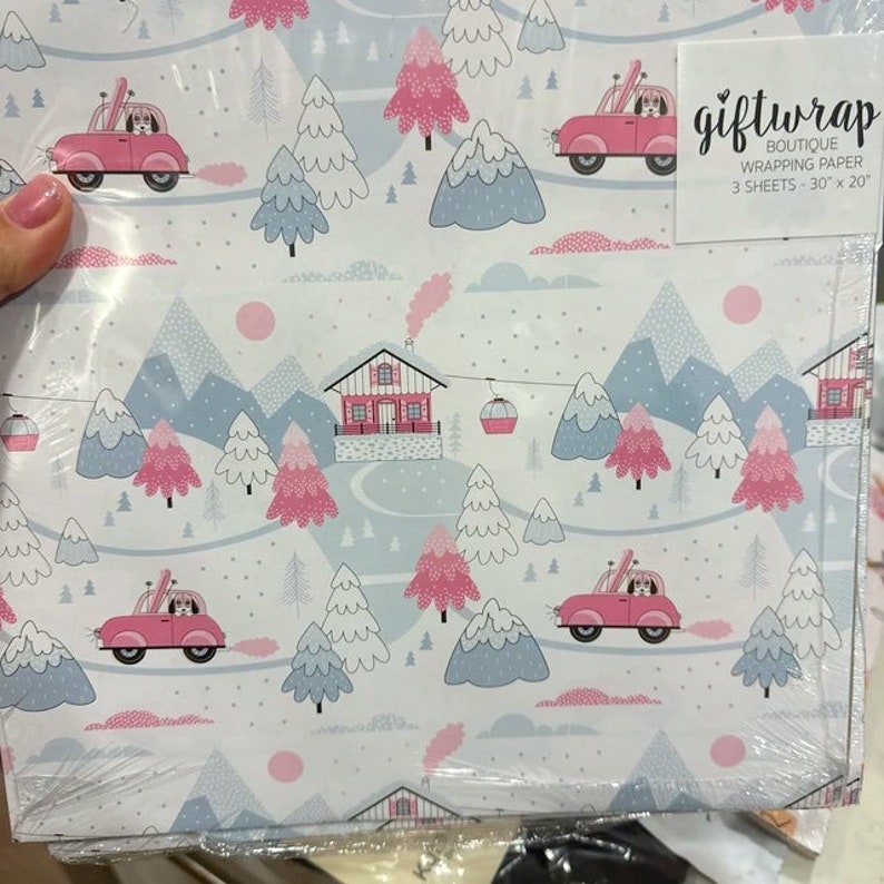 May include: A pack of three sheets of gift wrap featuring a winter wonderland scene with pink and blue houses, trees, and mountains. The wrapping paper is decorated with cars with dogs in them and the text "giftwrap boutique wrapping paper 3 sheets - 30" x 20".