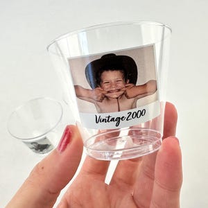 May include: Clear plastic shot glasses with a photo of a child wearing a hat and making a funny face. The photo is labeled "Vintage 2000". One glass is held in a hand, and another is in the background.